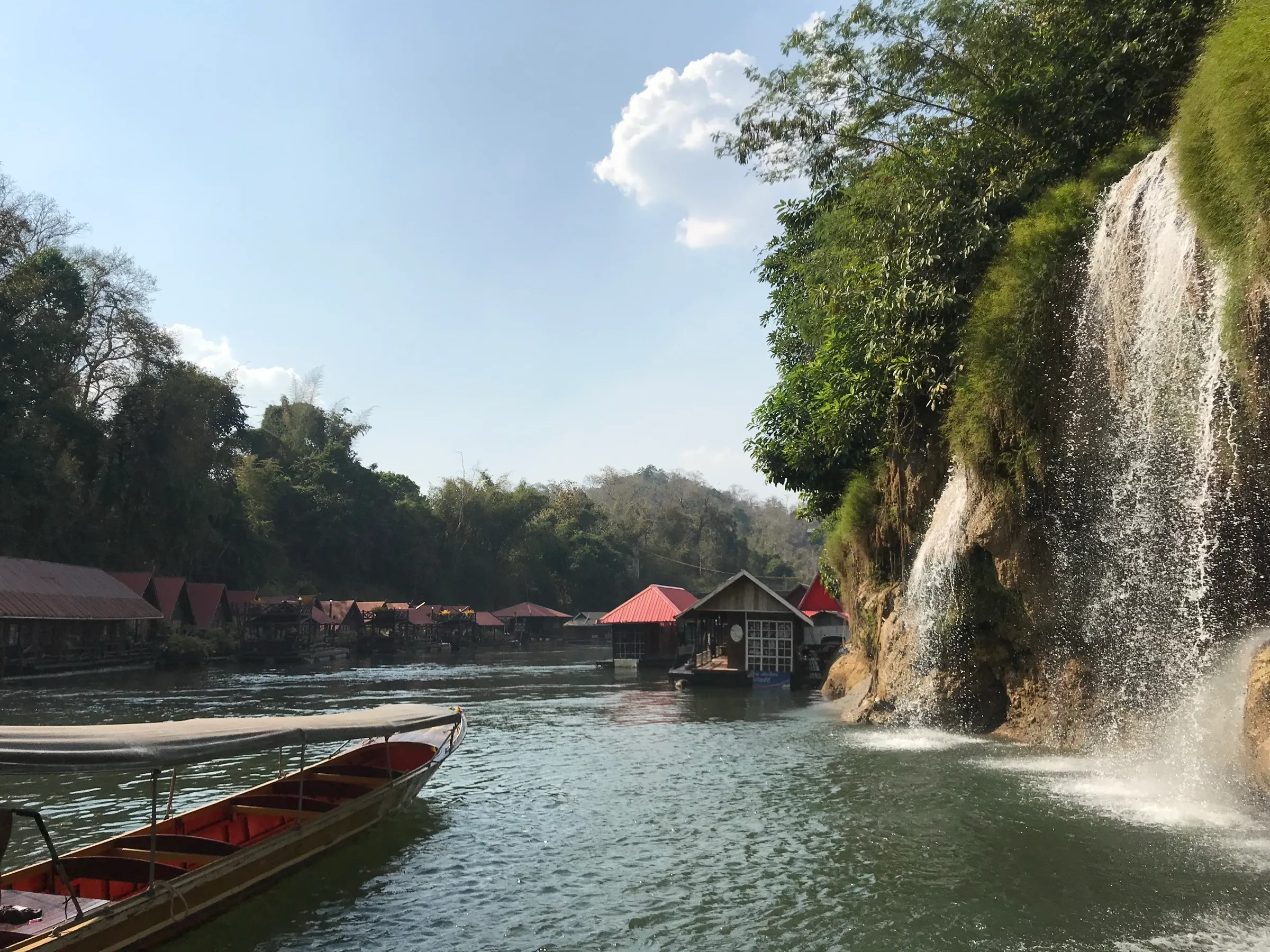 Wooden raft houses and a waterfall on the River Kwai at Sai Yok, a longtail boat moored in the foreground.