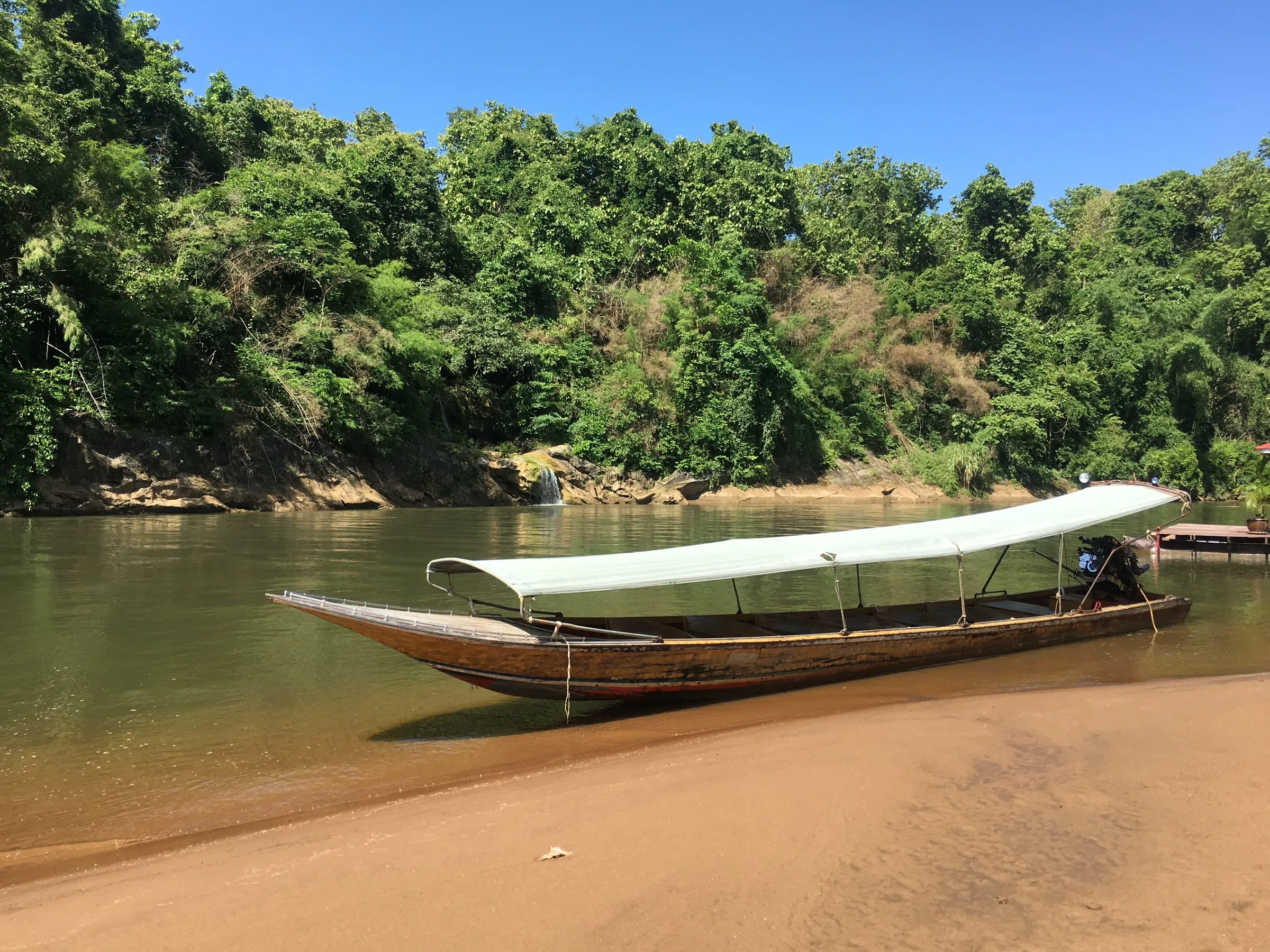 A wooden longtail boat beached on a sandbank below the forest at Sai Yok.