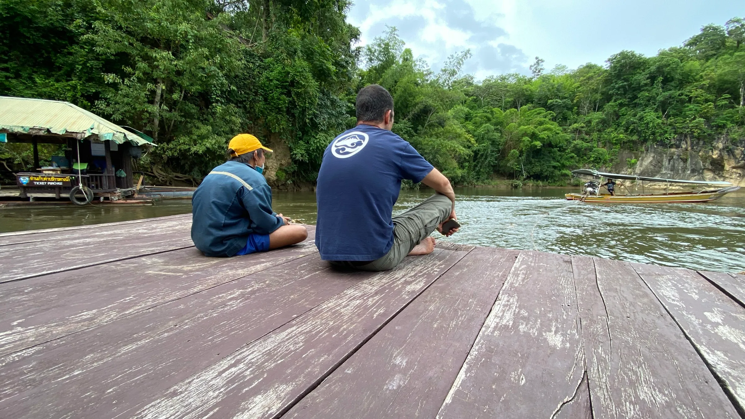 Two travellers sitting on the wooden deck of a floating raft house, looking out over the River Kwai.