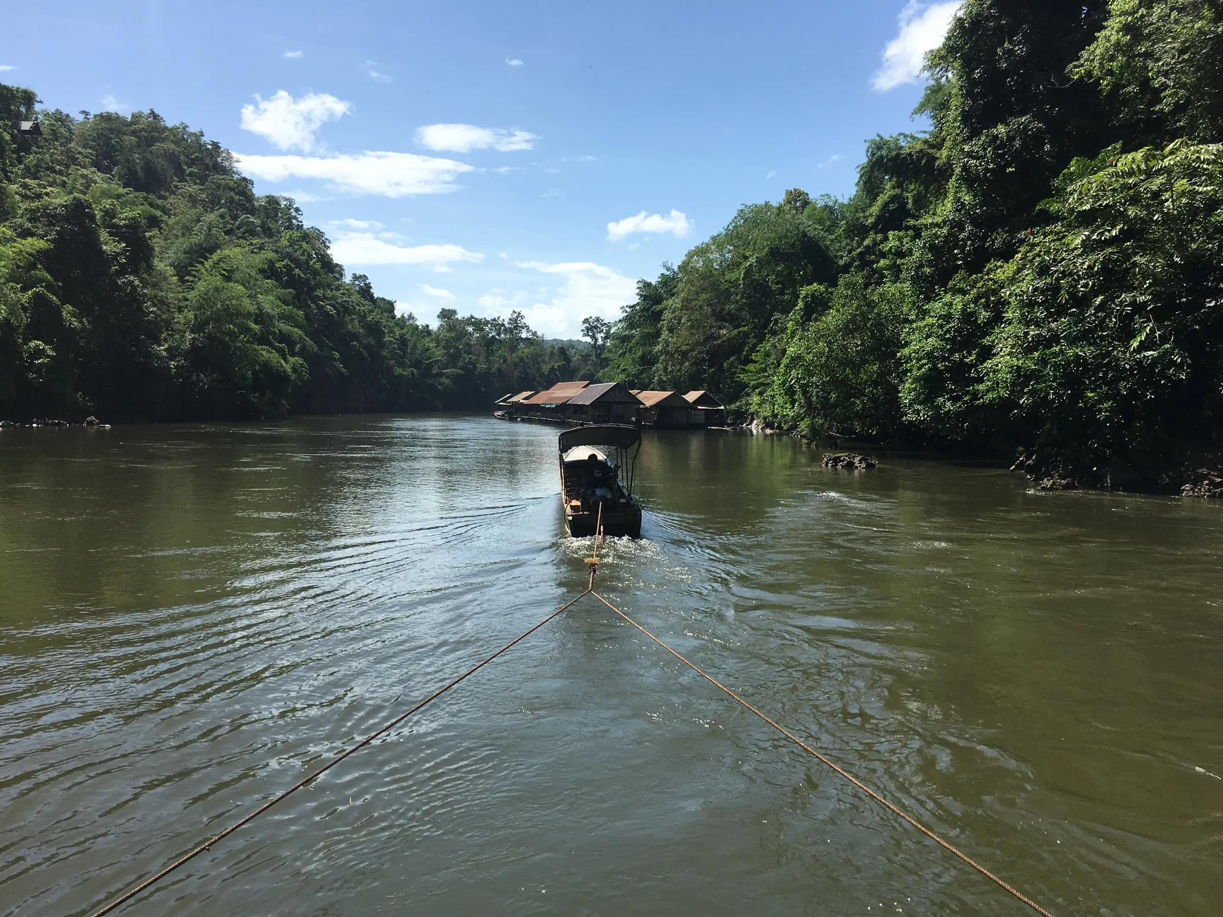 A longtail boat towing a wooden raft up the green River Kwai through the jungle.