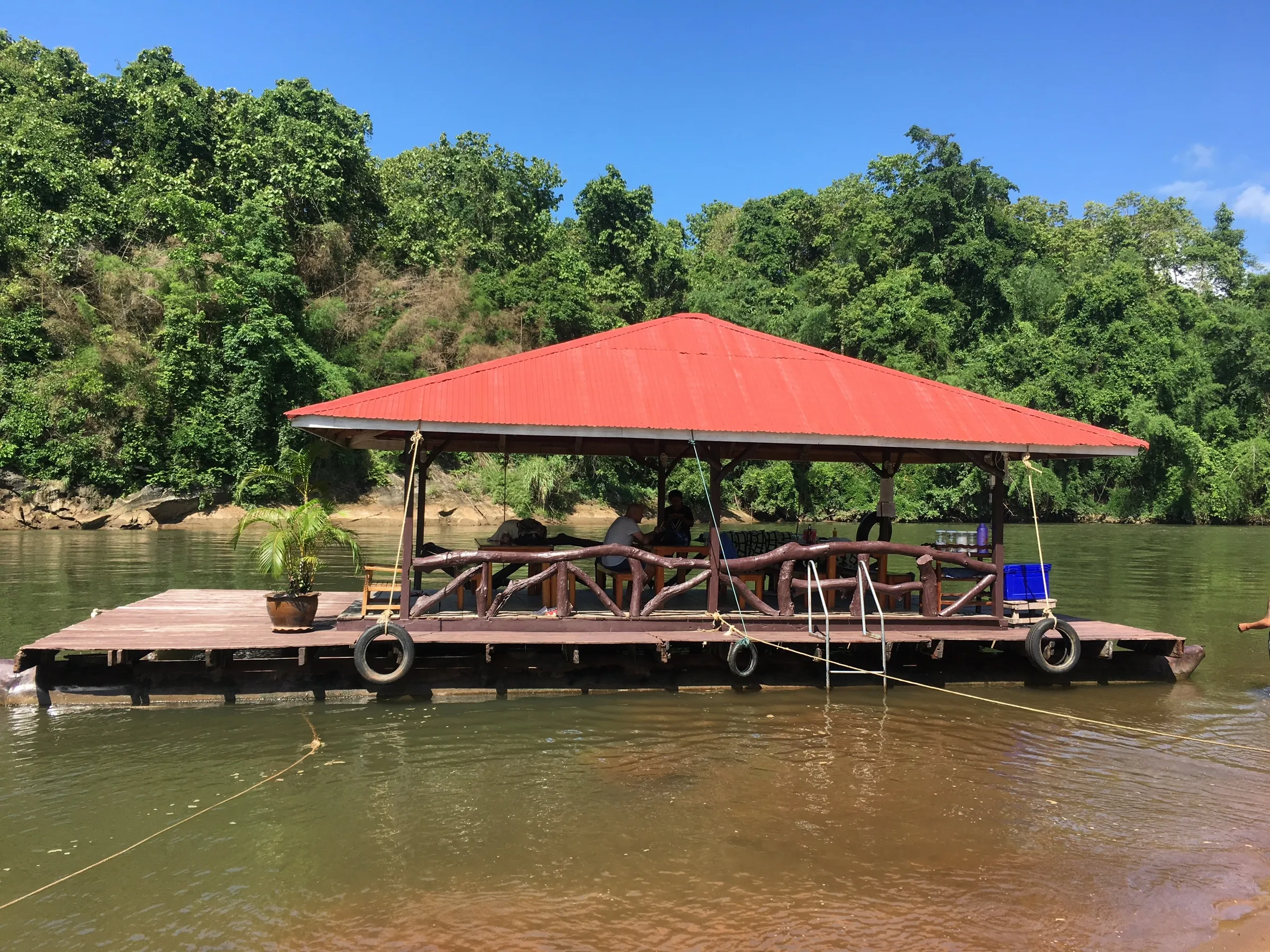 A floating raft with a red roof moored on the River Kwai, jungle behind.
