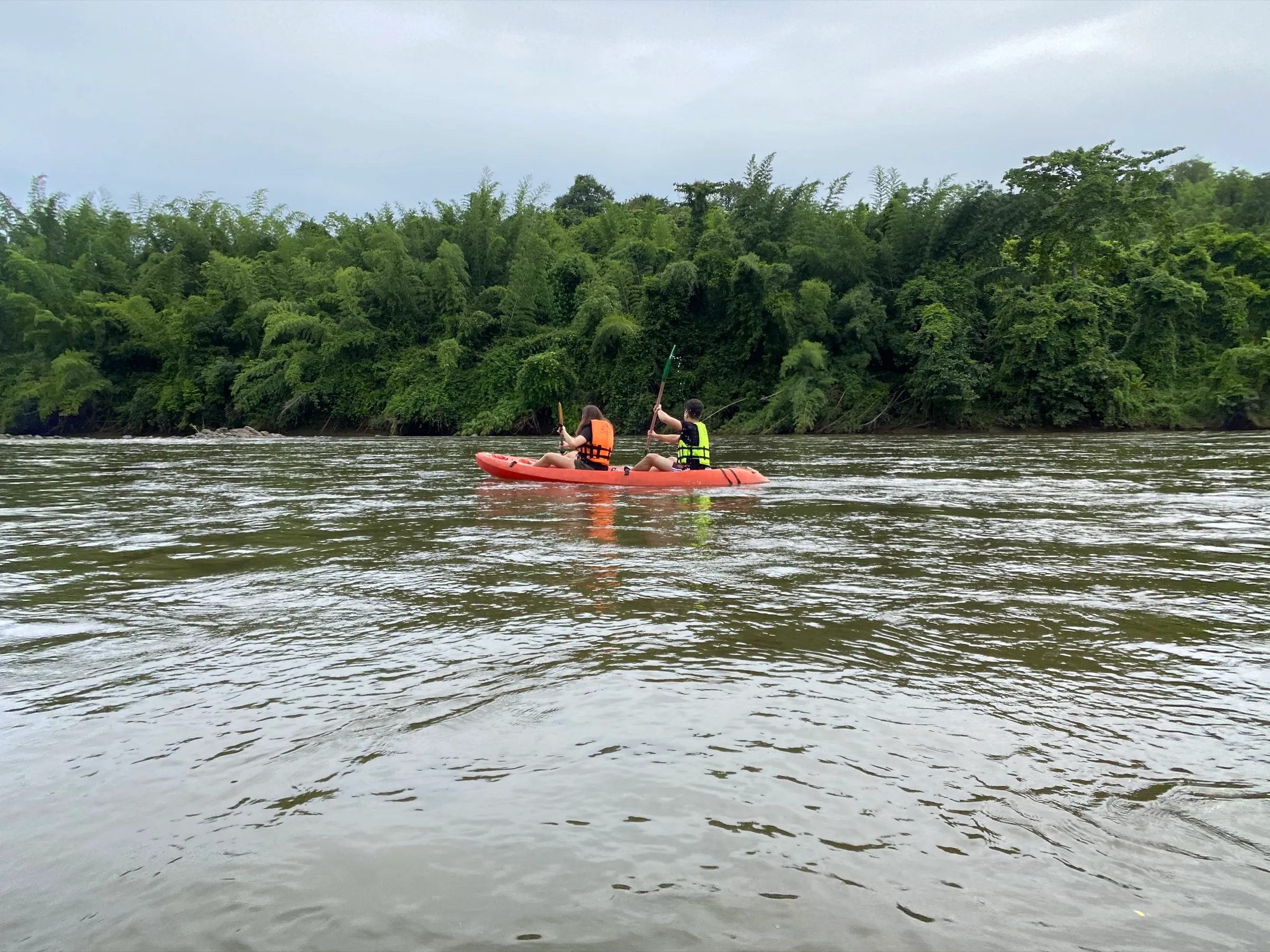 Two people paddling an orange kayak down the River Kwai past the forest.
