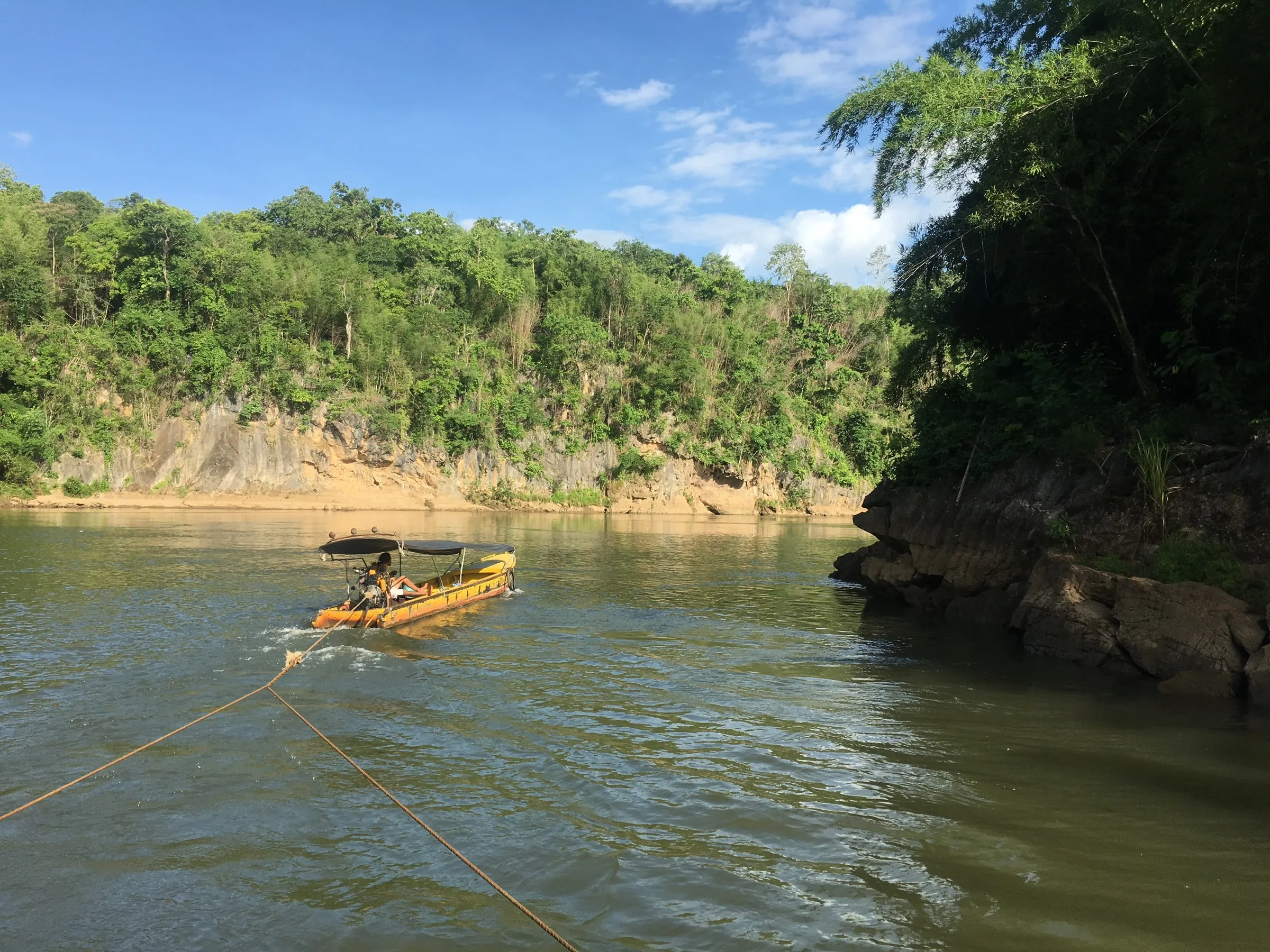 A longtail boat towing a raft on a rope down the River Kwai through the forest.