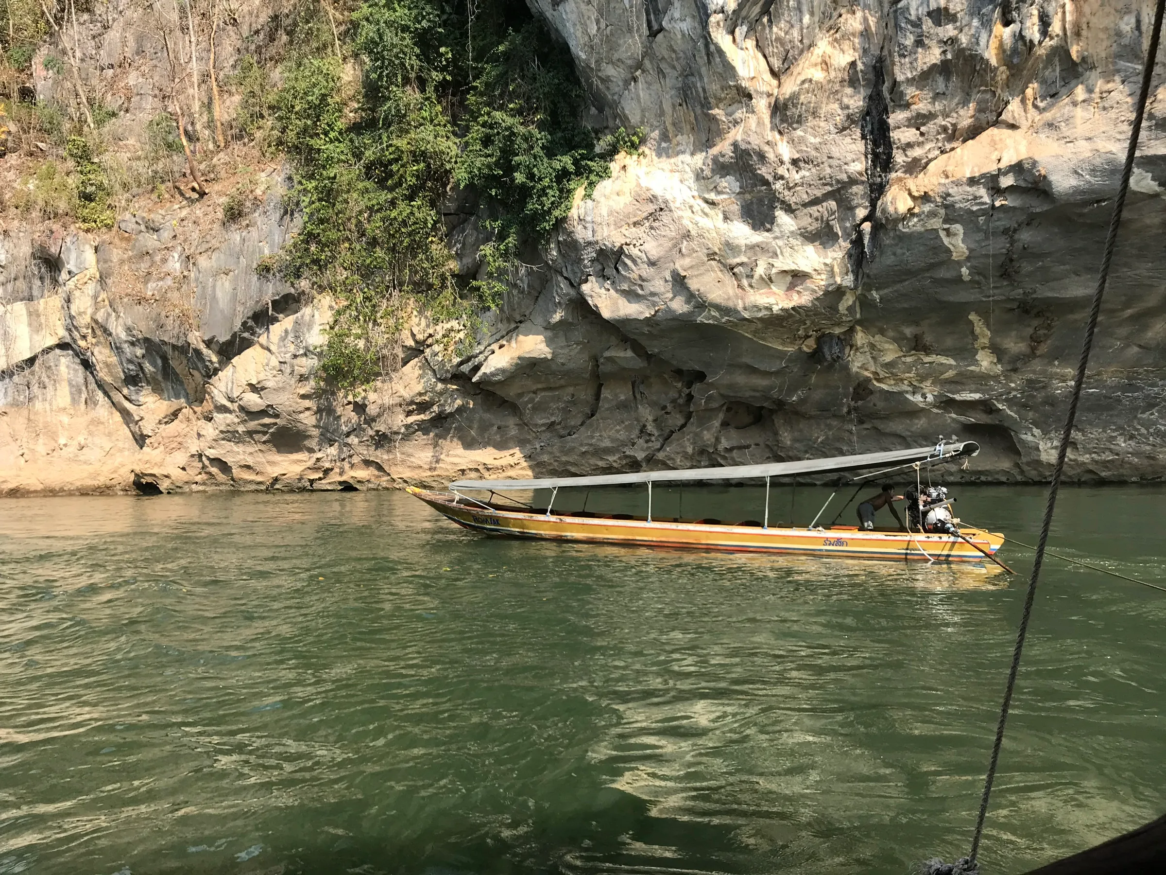 A longtail boat moored against a tall limestone cliff on the River Kwai.