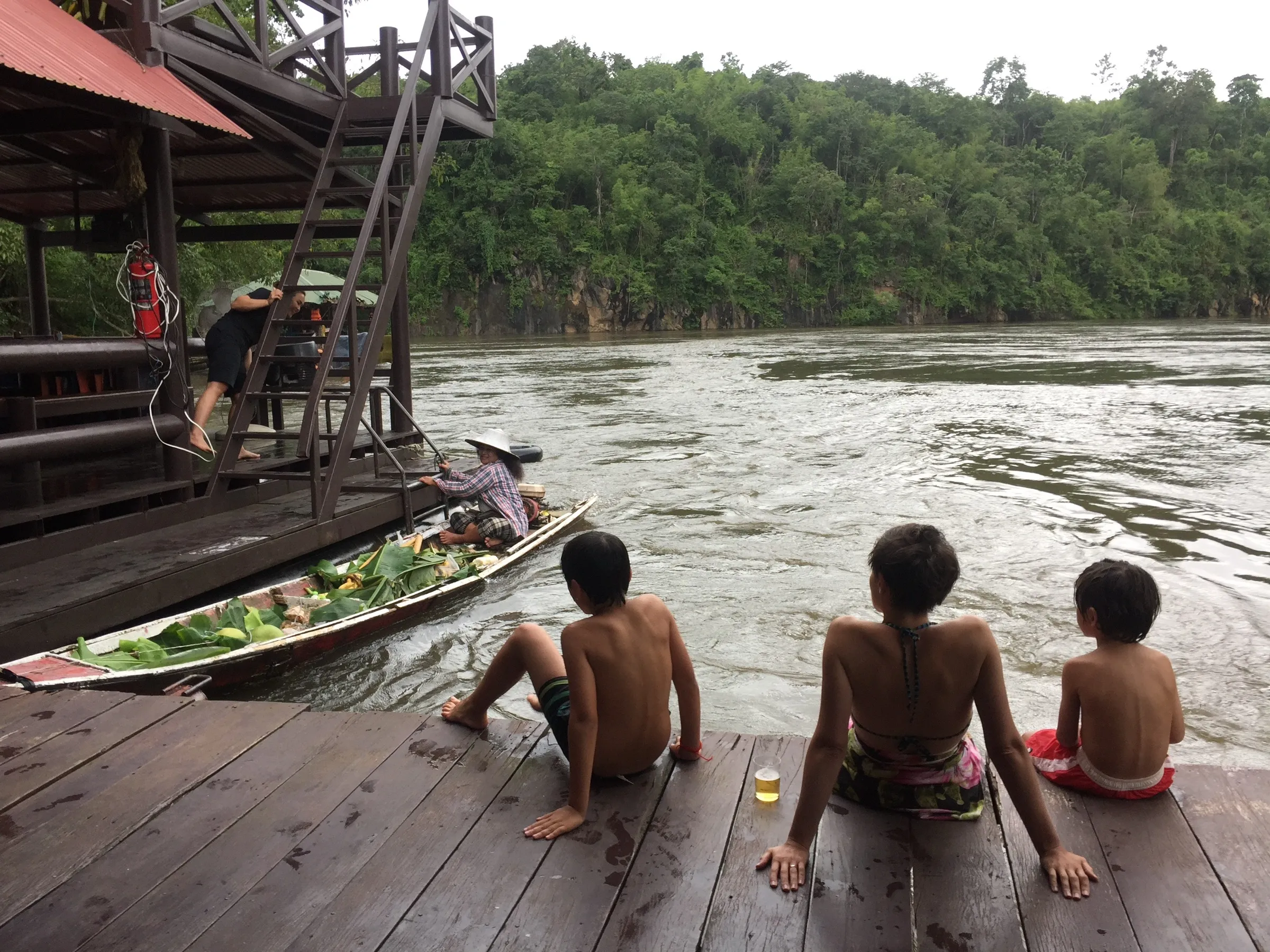 Children sitting on a raft deck watching a vendor’s food boat approach on the river.
